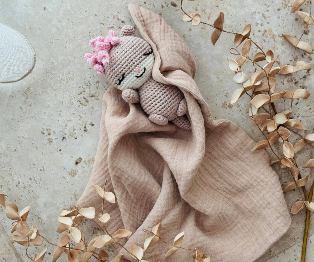 Pink knitted  otter toy with pink flowers draped over a beige blanket on a textured surface with dried leaves.