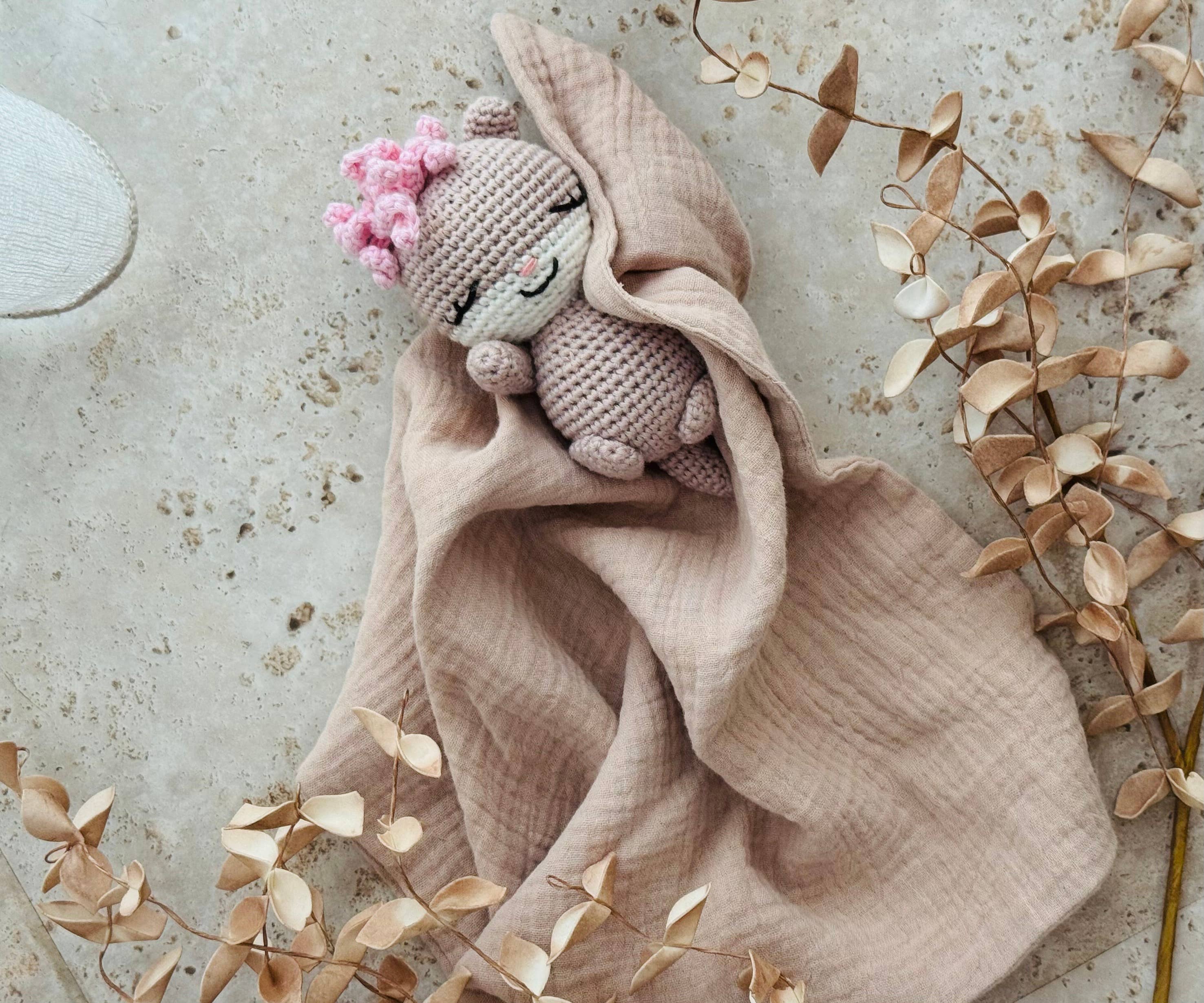 Pink knitted  otter toy with pink flowers draped over a beige blanket on a textured surface with dried leaves.