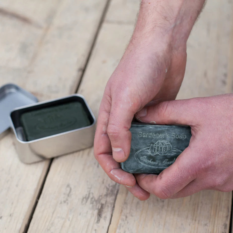 Person holding a bar of soap with 'Gardener's Soap' branding on a wooden surface.