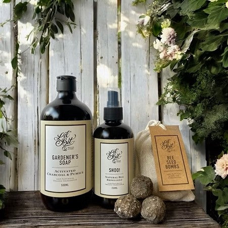Two bottles of 'Lil' Bit' garden care products on a wooden surface with plants and stones in the background.