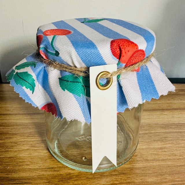 Glass jar wrapped in a colorful striped cloth with a cherry design, tied with twine and a white tag.