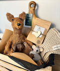 Gift basket with plush toys, wooden cutting board, and baby blanket on a white surface