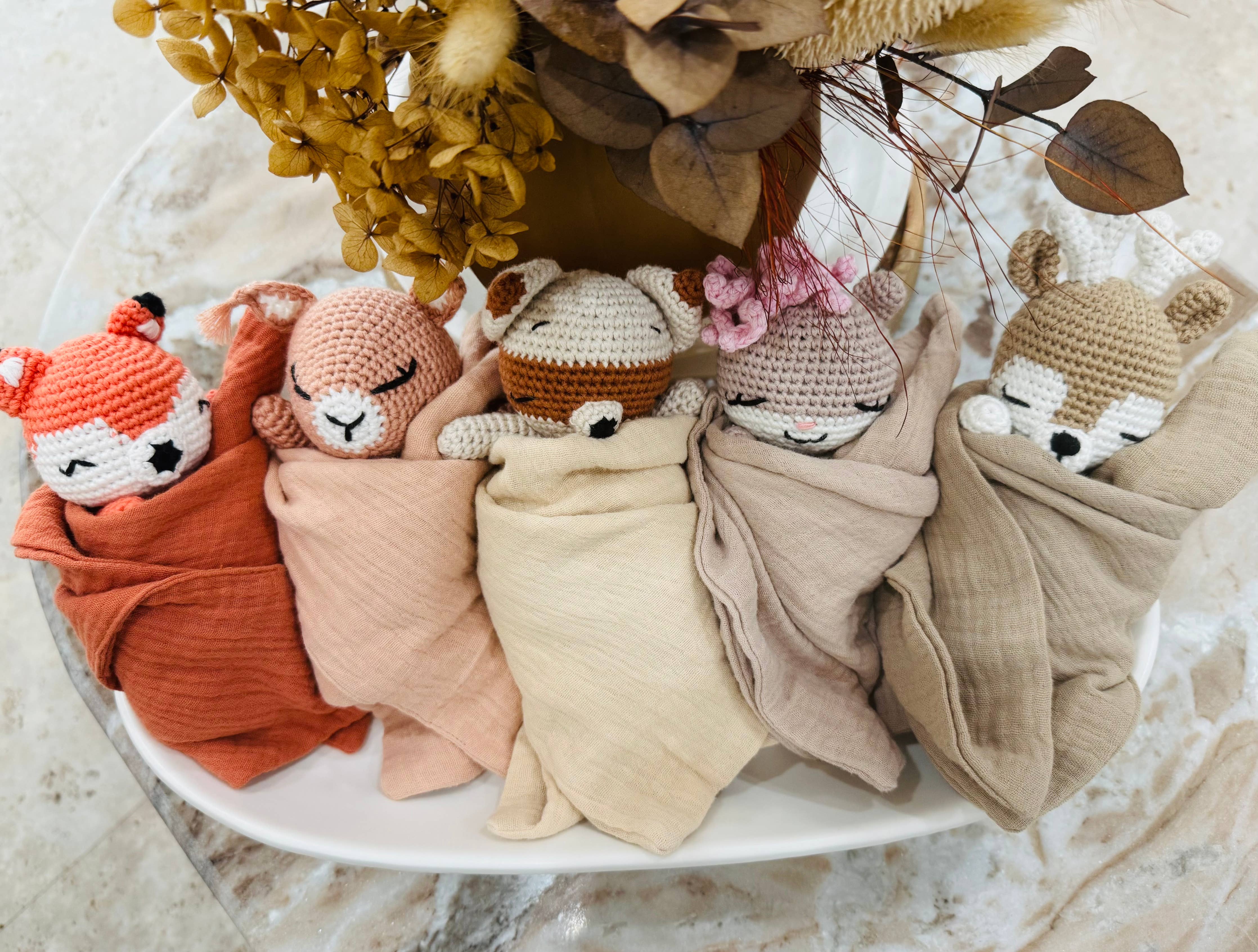 Five knitted animal toys wrapped in colorful blankets on a marble surface with decorative elements.
