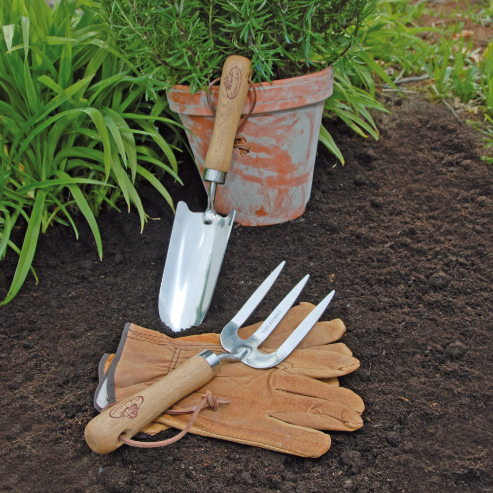 Gardening tools including a trowel, fork, and gloves on garden soil with a potted plant in the background.
