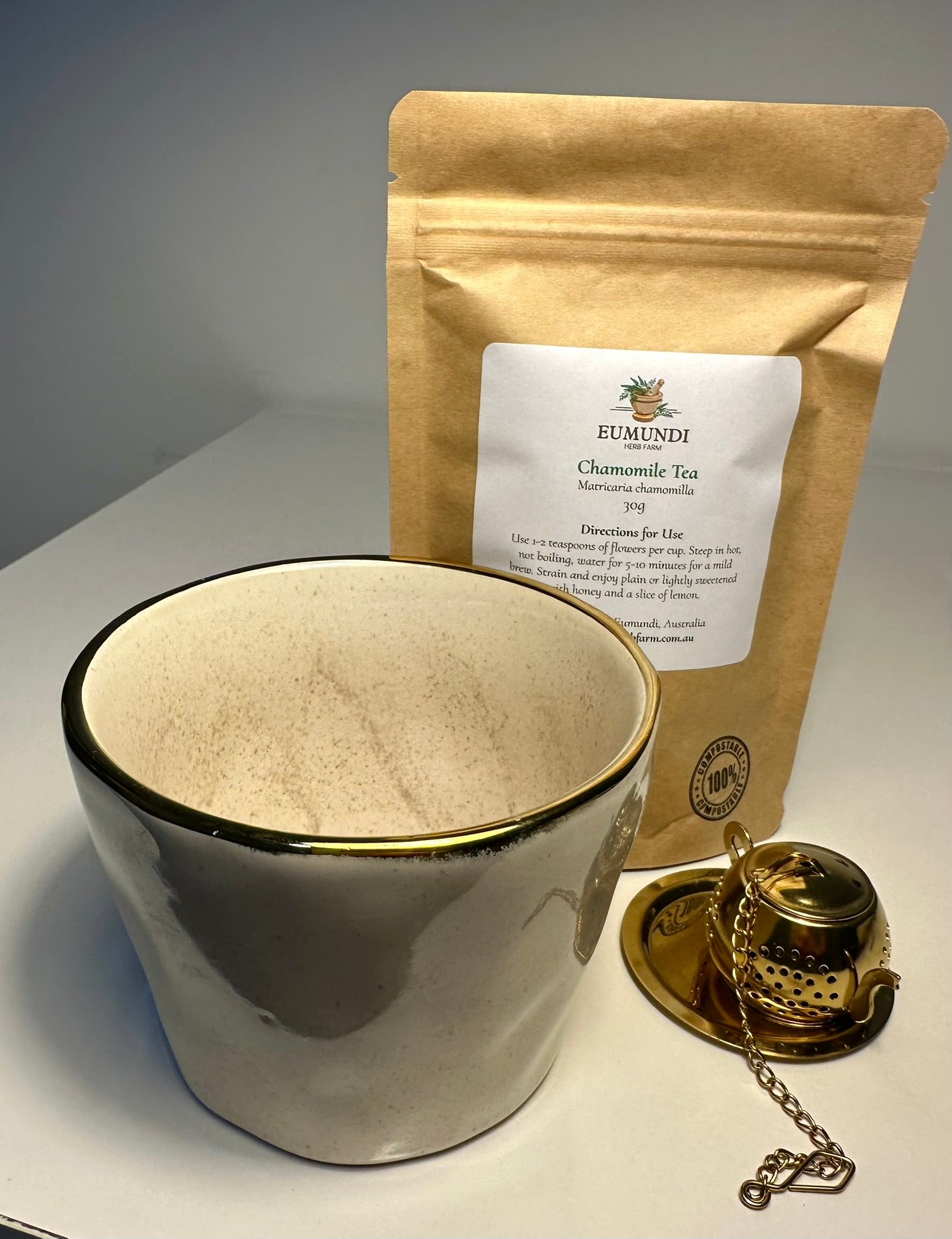 White ceramic tea cup with gold rim, Eumundi chamomile tea package, and gold tea infuser on a gray surface.