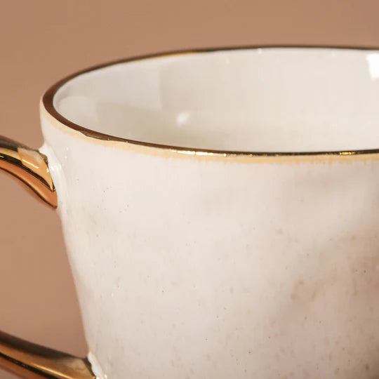 Close-up of a white ceramic cup with gold rim and handle on a matching saucer.