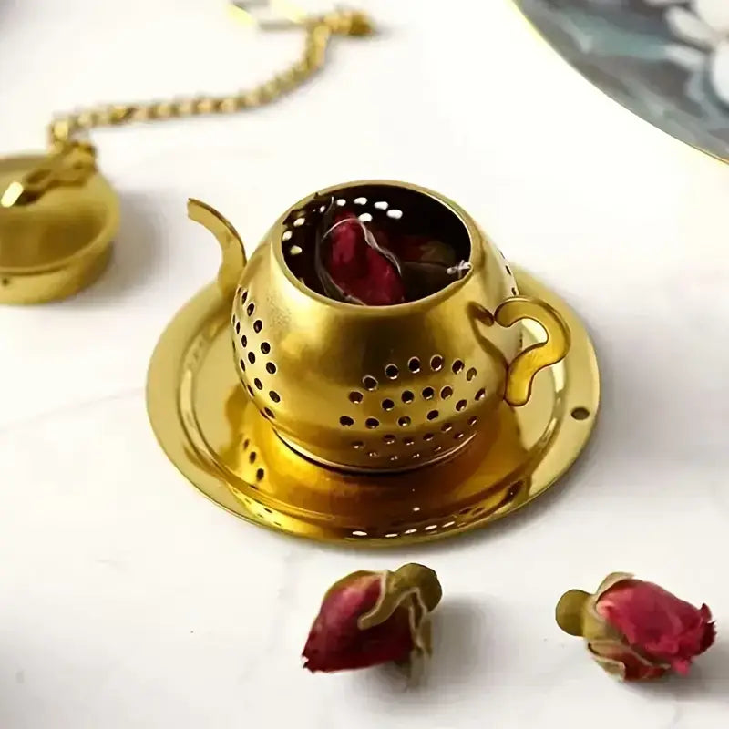 Gold tea infuser with teacup design on a white surface, surrounded by dried rose petals.