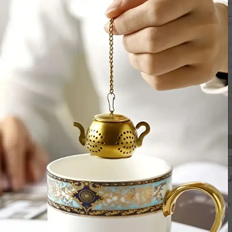 Gold tea infuser being held over a decorative teacup.