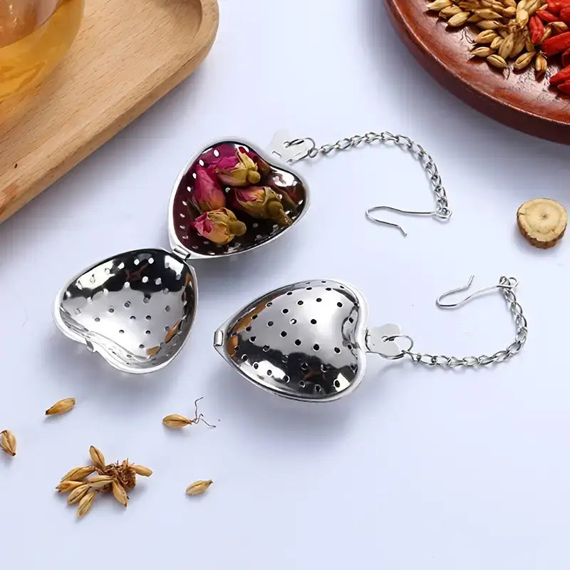 Heart-shaped metal tea infusers with a chain on a white surface with dried herbs.