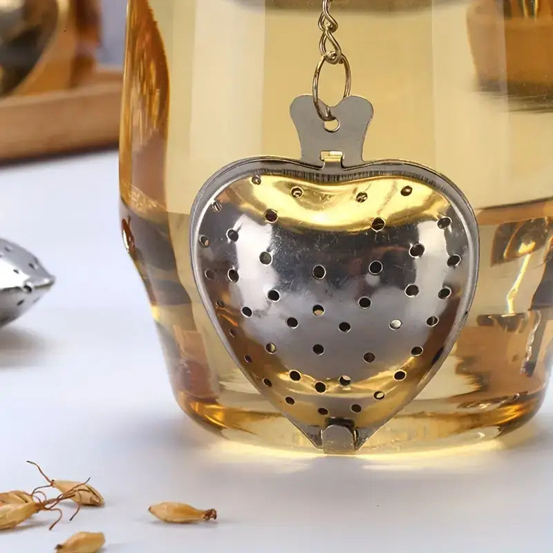 Heart-shaped metal tea infuser in a glass of tea with loose tea leaves on a white surface.