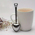 Heart-shaped tea infuser in front of a white mug with tea, on a light background.