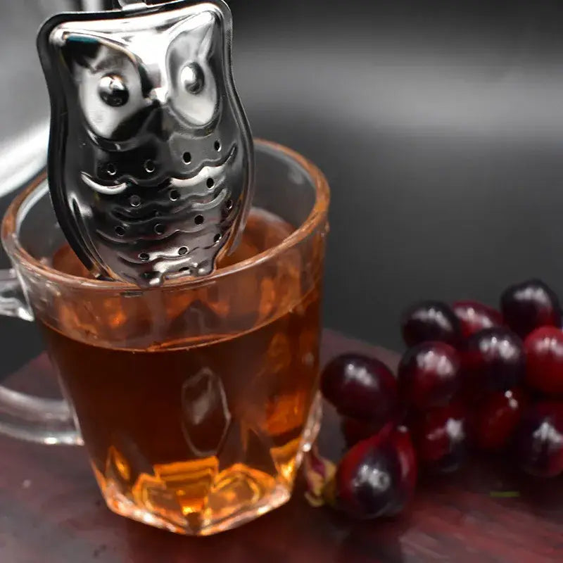 Tea infuser shaped like an owl in a glass of tea with grapes on a dark surface