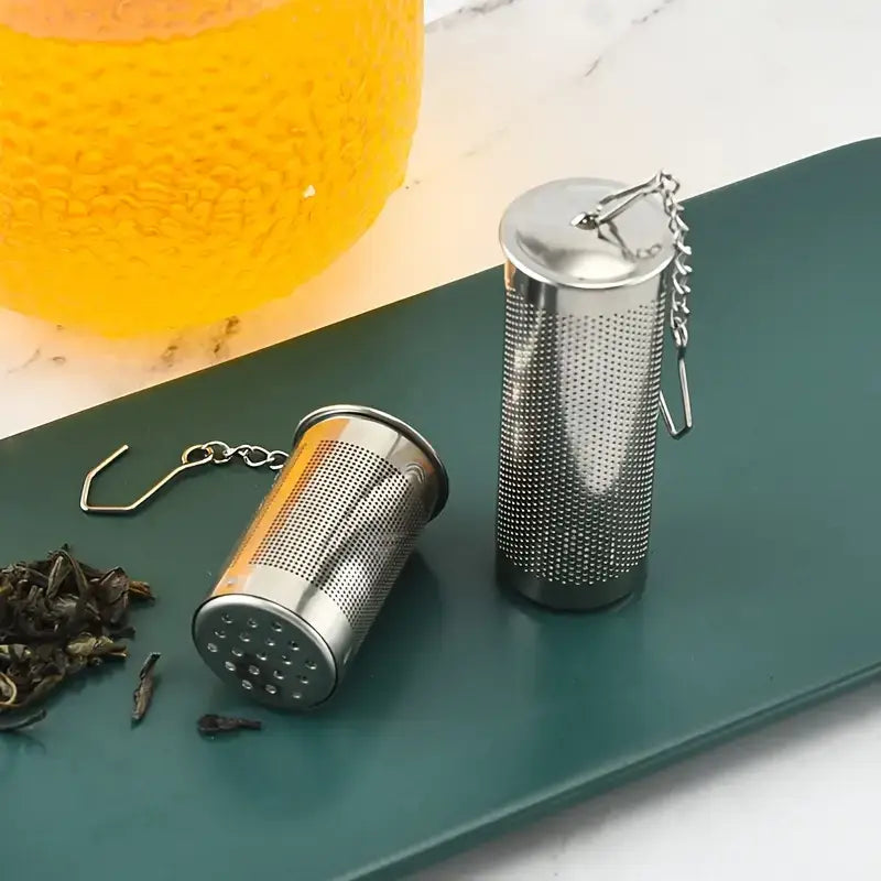 Metal tea infusers on a green tray with dried tea leaves and a glass of tea.